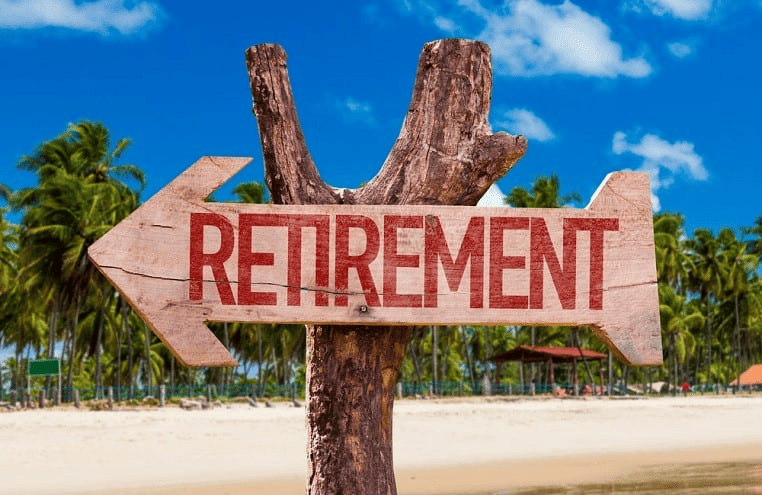 Image of a arrow-shaped sign with the word "Retirement" on it nailed to a tree trunk. Background features a beach with tropical palm trees.