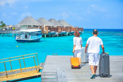 image depicts a couple standing on the wharf of a tropical island with their luggage. Villas over the water and a boat are in the background
